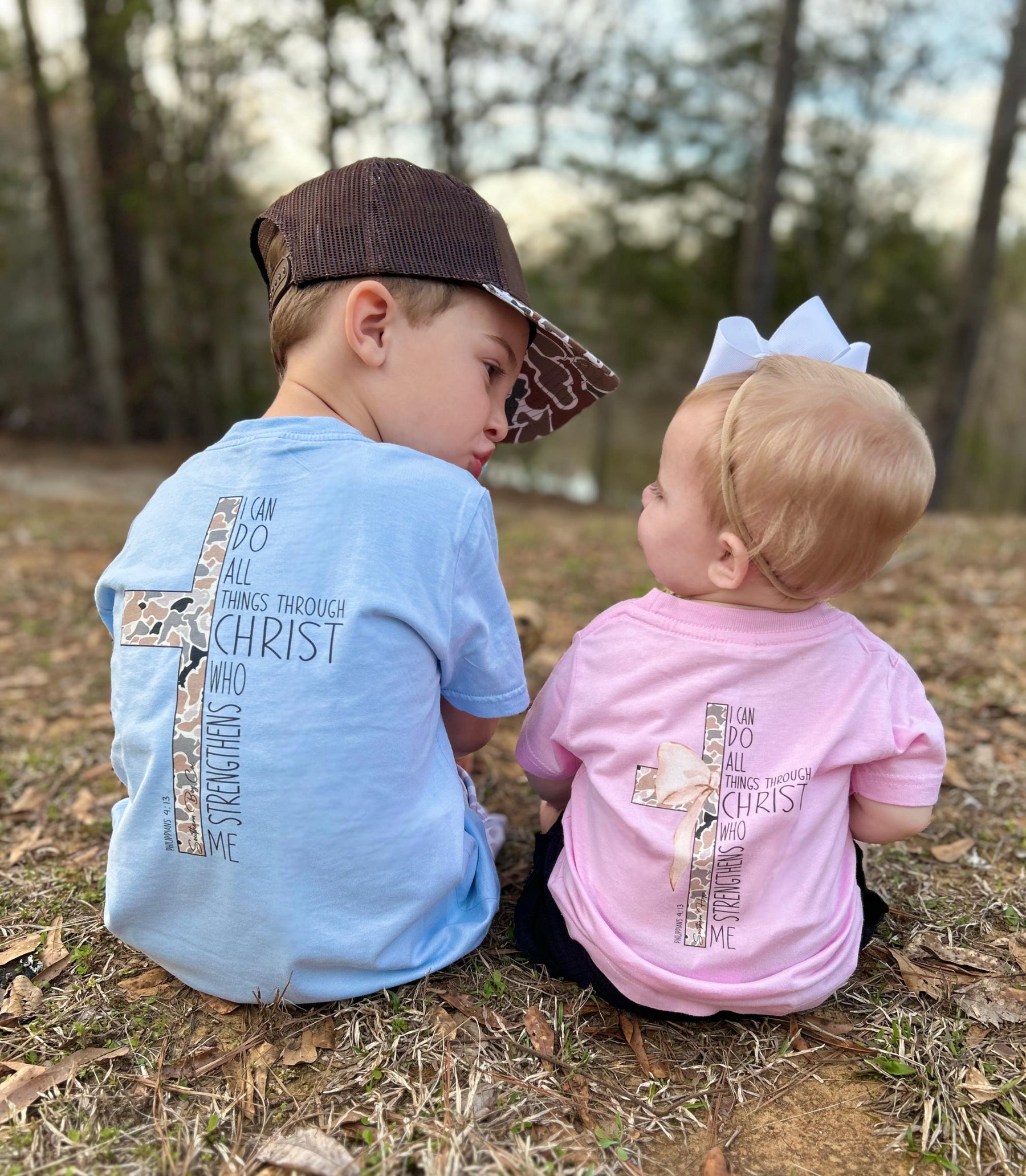 Young Southern boy and toddler girl sitting outdoors wearing faith-based t-shirts with camo cross designs and the Bible verse Philippians 4:13, 